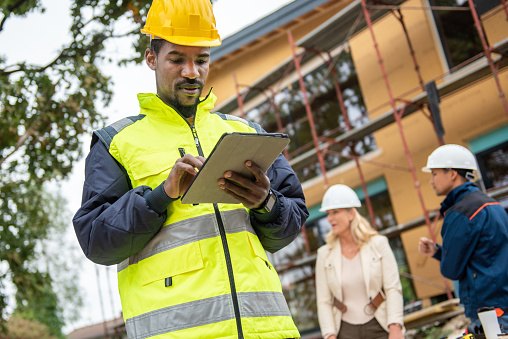 African american building contractor in protective hardhat and waistcoat using digital tablet while standing at construction site outdoors.