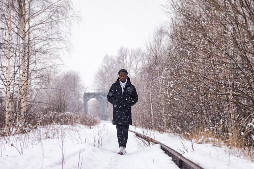 African american man walking on railway track in snowy russian winter in countryside forest outside, falling snow. Positive black man going outdoors. Leisure activity concept. Copy text space for ad