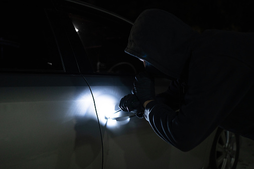 Asian man thief in black mask trying to open car using custom tool and flashlight. Car robber.
