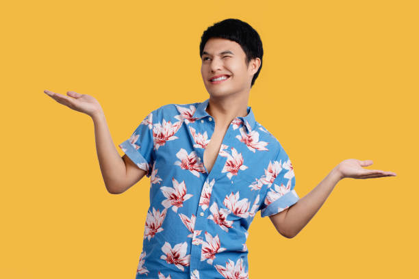 istock Smiling young handsome Asian man  presenting with open palms gesture on yellow background in studio. He in blue floral shirt for Songkran festival in Thailand. 1472467416