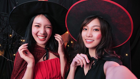 Happy two Asian women in Halloween costumes smiling and taking selfie at house party, Funny friends in costumes at night Halloween party