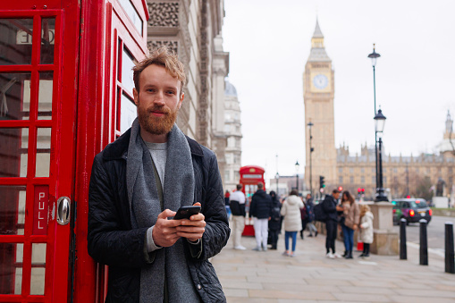 Man with a smartphone in his hands standing near a red telephone booth against the backdrop of the Big Ben tower