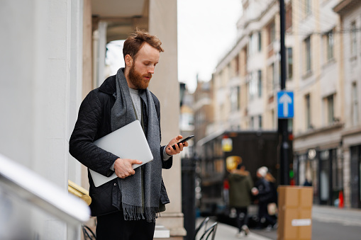 Bearded business man in a jacket and scarf with a laptop using a smartphone while standing on the street near the entrance in the building