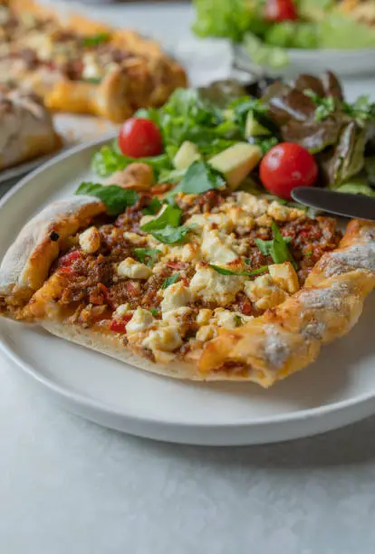 Delicious turkish pide or turkish pizza with spicy ground beef, tomatos, red peppers, chili, onions, garlic and parsley. Topped with feta cheese. Served hot and ready to eat on a kitchen table background. Closeup, front view