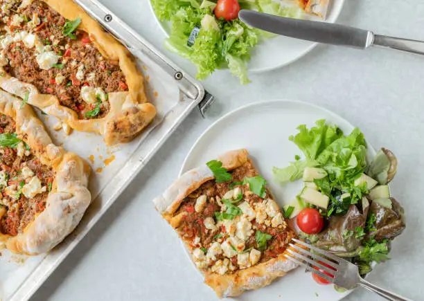 Homemade fresh baked turkish pide or turkish pizza with bell peppers, chili, onions, tomatoes and herbs. Baked with feta cheese topping and served ready to eat on white kitchen table background with plate and baking sheet. Flat lay