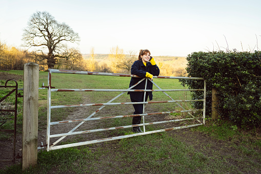 Full length view of woman wearing warm winter clothing, leaning against pasture gate, and smiling while conversing with caller.