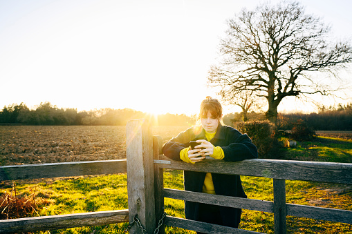 Front view lens flare of early 20s Caucasian woman leaning against wooden fence, scrolling portable device, agricultural field and setting sun in background.