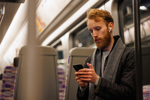 Male subway passenger sits on a train listening to music on headphones and using a smartphone