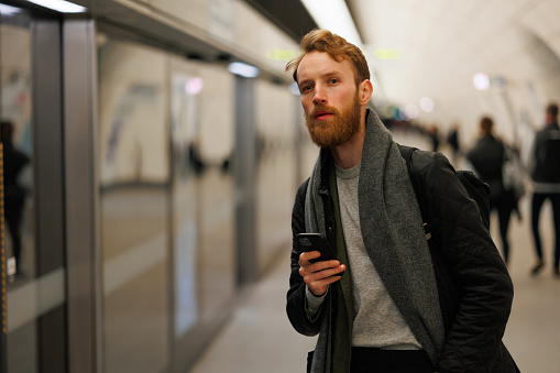 Bearded man is standing on the platform of a subway station with a smartphone in his hands, looking at the train