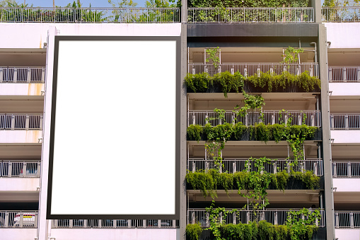 Blank white vertical advertising billboard banner mockup, outside multi-storey carpark with lush green plants. Large digital display screen poster, an out-of-home OOH media display space.