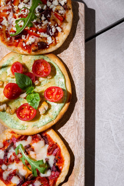 istock Three mini pizzas with tomatoes, cheese, shrimps, arugula, ketchup, mozzarella, pepper and olives lying on a wooden board that stands on a concrete table 1472498903