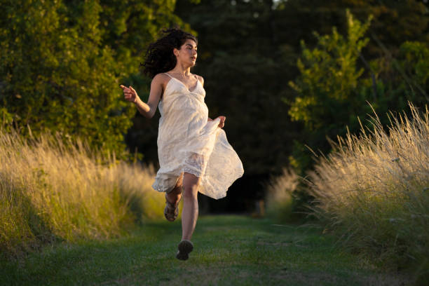 A shot of a female running through a grassy meadow during golden hour near Toulouse, France. She is wearing a white dress and looking into the distance as she is running.