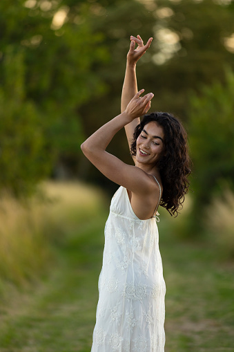 A waist-up portrait shot of a female standing with her arms raised above her head as she dances in a meadow at a golden hour near Toulouse, France. She is wearing a sleeveless white dress and is looking over her shoulder. The shot has a shallow depth of field with soft bokeh.