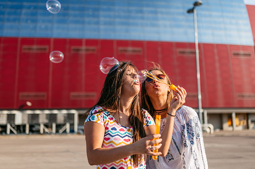 Two beautiful young female friends blowing soap bubbles in the parking lot.