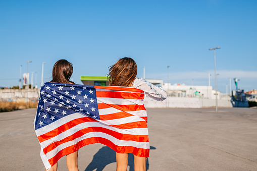 Two beautiful young women wrapped in USA flag in the parking lot in the summer.