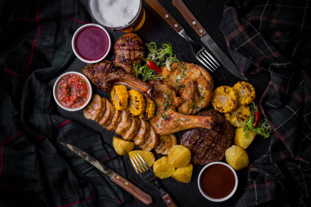 istock Grilled beef steak on the bone chicken leg, pork steak, meatloaf with potatoes, corn, different types of sauces, salad with cutlery, and a glass of beer on a stone board. Horizontal orientation 1472507497