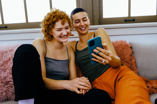 Two cheerful young women sitting together on sofa, smiling and enjoying each others company while using their .smartphone to stay connected reading text.
