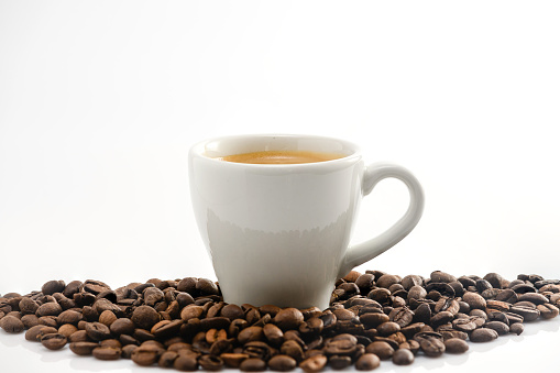 A cup of espresso in a pile of roasted coffee beans on a white background.
