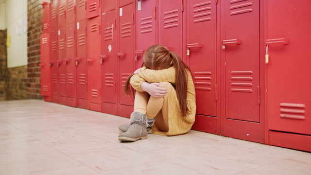 Sad girl, student and depression sitting on floor in stress, trouble or bullied at school hallway. Lonely female child learner suffering anxiety, problems or mental health issues against lockers