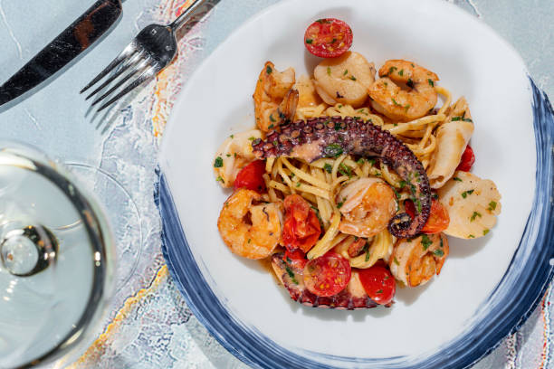 Pasta with seafood. Homemade pasta with scallop, octopus, shrimp and cherry tomatoes in a creamy herb sauce. Pasta lies in a round, light ceramic plate. The plate stands on a light marble table. Nearby is a glass of prosecco, cutlery lies. pasta con marisco. pasta casera con vieira, pulpo, camarones y tomates cherry en una cremosa salsa de hierbas. la pasta se encuentra en un plato de cerámica redondo y ligero. el plato se encuentra sobre una mesa de mármol claro. cerca hay un vaso de pros