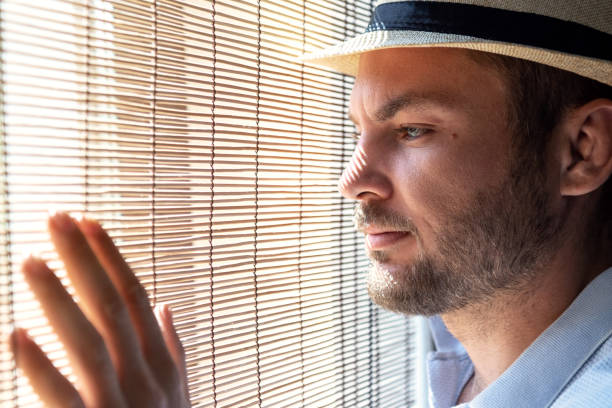 istock Man in straw hat looking out the window through straw blinds. 1472512487