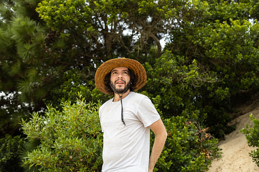 Tranquil bearded young man with curly hair wearing a straw hat and white shirt looking away standing on a dune with trees on background