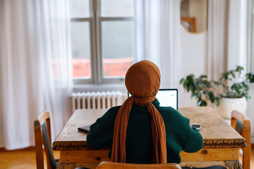 High angle view of woman wearing hijab sitting at work desk, using laptop in home office