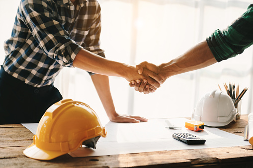 Construction team shake hands greeting start new project plan behind yellow helmet on desk in office to consults about their building project. in sun light