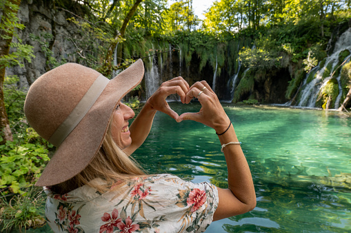 She makes a heart shape frame with hands on beautiful lake and waterfalls in Croatia.