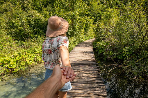 They travel in Croatia and discover national parks in the country. Man's first perspective following girlfriend ahead. They walk on wooden footbridge near lake and waterfalls