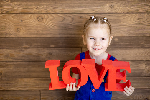 a small child girl with a red inscription Love in her hands smiles or laughs against a wooden wall, the concept of Valentine's day, an empty space for text