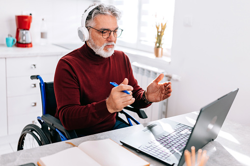 A mature Caucasian man in a wheelchair is gesturing while talking with a client in a video call.