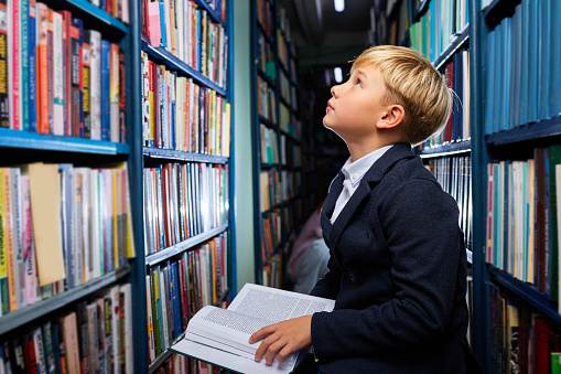 school boy sits in library looking up at books shelves, in search of books.