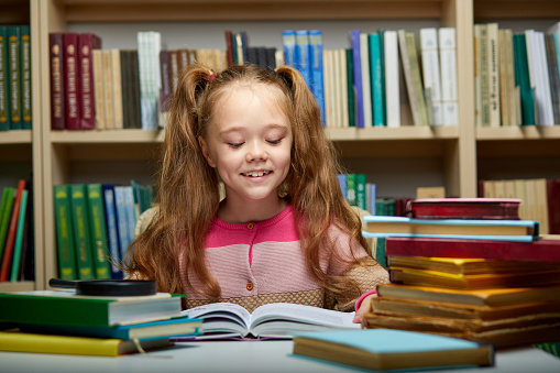 preschooler little girl reading a book in the library,little caucasian girl sits with books near a bookcase