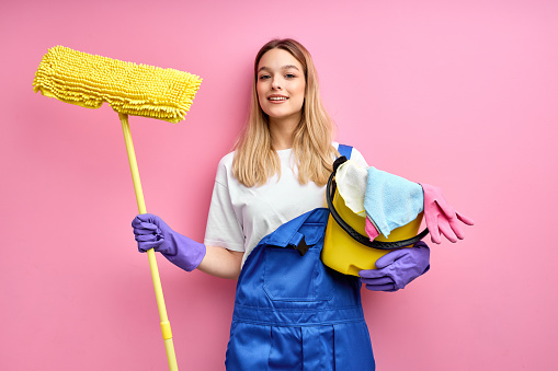 Smiling nice caucasian woman in blue coveralls and rubber gloves look at camera holding cleaning supplies. isolated pink background, portrait of pretty female ready to clean