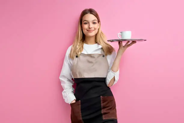 nice woman waitress in apron, offering cup of delicious tasty coffee, stand smiling, friendly staff of restaurant. isolated over pink studio background