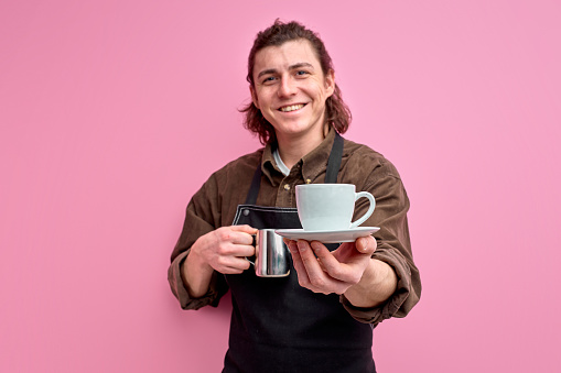 Male waiter offering cup of coffee isolated on pink background, smiling man in apron give mug of beverage to camera. focus on cup