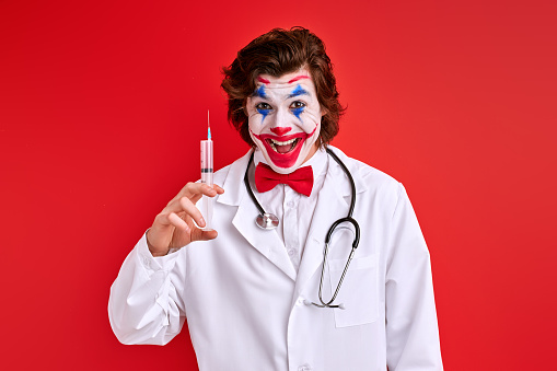 male clown pediatrician prepared a syringe isolated over red background, going to treat someone.