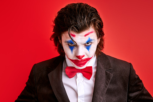 portrait of virile harsh illusionist with colourful make-up confidently looking at camera, magician in suit isolated over red background