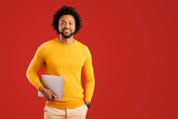 Curly young African-American male entrepreneur stands in studio isolated on red and carrying trendy laptop. Cheerful black guy in casual clothes looks at camera stock photo