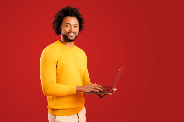 istock Optimistic african-american male student using laptop pc isolated on red background, smiling multiracial freelancer man looking enjoying remote work, advertising 1472539937