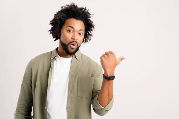istock Cheerful overjoyed African-American guy points fingers at empty copy space, isolated on white background. Multiracial man recommends deal, pays yours attention, presenting new product 1472539976