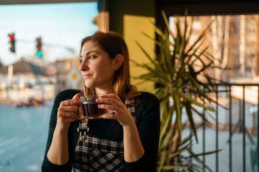 Young woman looks out the window while sitting in a coffee shop and drinking a hot tea.