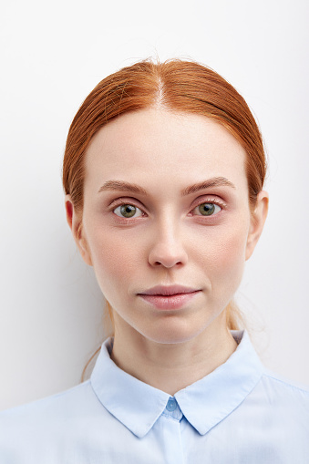 Close-up portrait of female with natural red hair in blue blouse posing at white studio wall, having calm expression looking at camera. Headshot for passport
