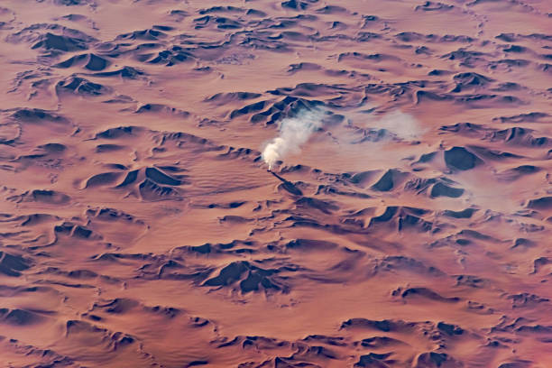 White gas smoke comes out of a hole in the middle of reddish sand dunes afar in the Sahara desert of Algeria. Aerial view from an airplane in a sunny day with sunlights and shadows