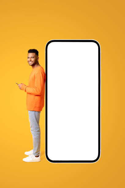 istock Smiling attractive guy standing near huge smartphone with empty blank screen, man using phone, advertising new app or website, mockup 1472546925