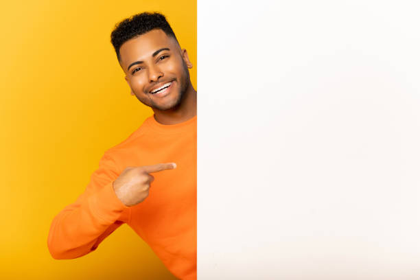 Look here. Smiling indian man peeking out from behind white empty paper poster, presenting novelty, arab guy paying attention at blank billboard banner isolated on yellow, copyspace, advertising