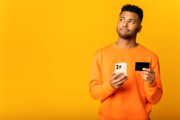 Thoughtful handsome indian guy holding credit card and smartphone, dreaming about long-awaited purchase standing isolated on yellow stock photo