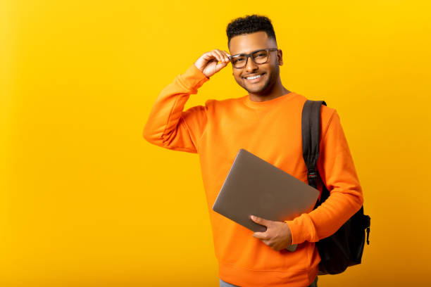 Smart inteligente indian male employee or freelancer man holding laptop and looking at camera with happy smile, isolated on yellow background stock photo