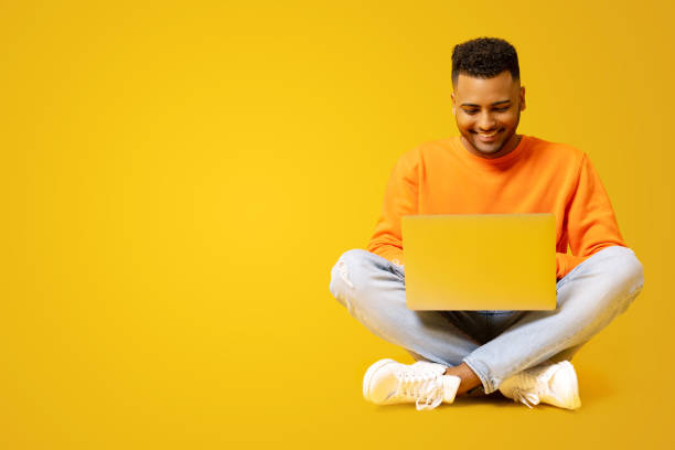 istock Serene Indian male student is using laptop for online studying sitting on the floor isolated on yellow, preparing homework, researching topic, positive hindu guy typing 1472547044